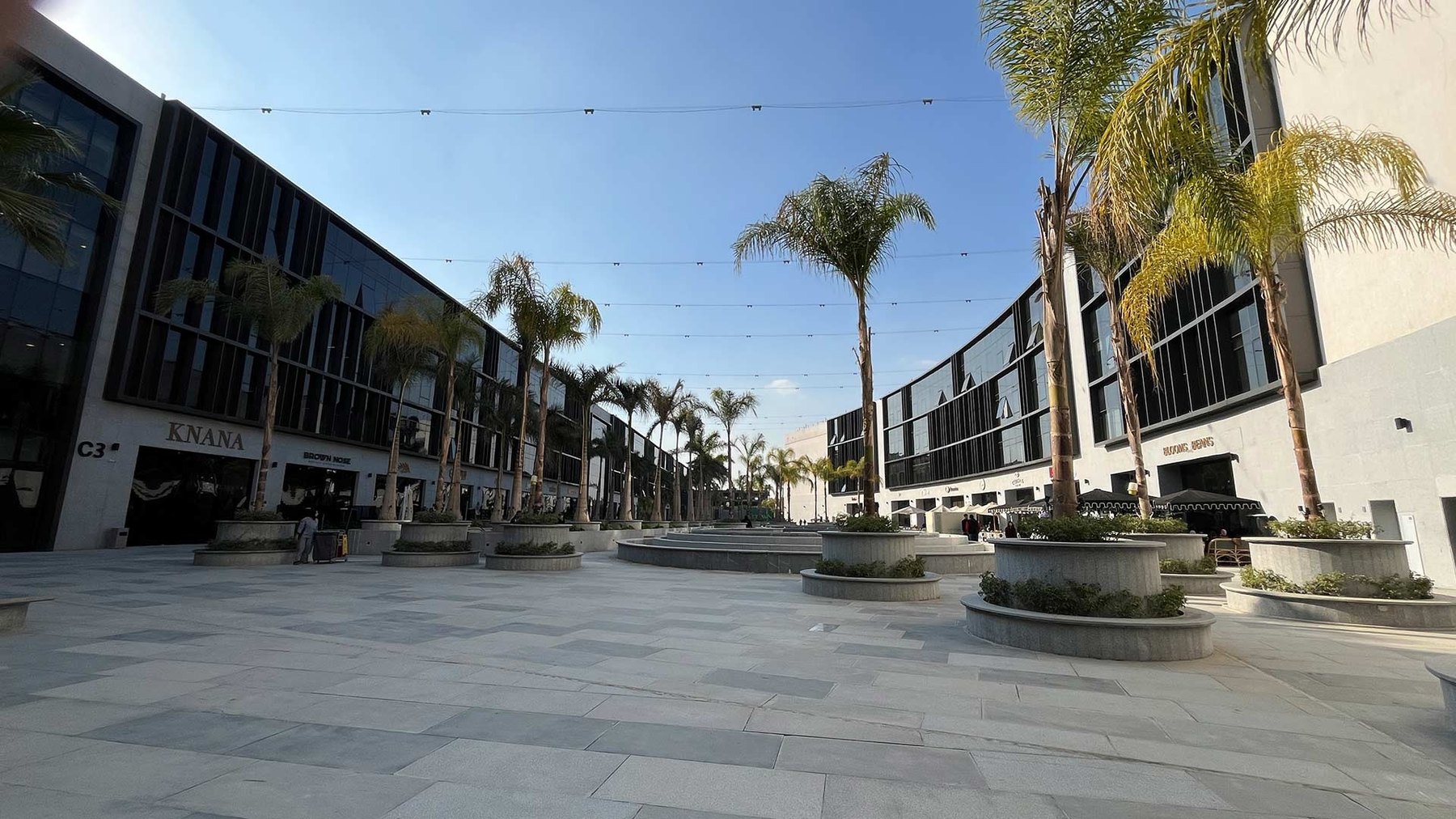 Wide stone plaza at Laketown, New Cairo, lined with tall palms in round planters between two three-story glass-and-stone commercial buildings under a clear sky