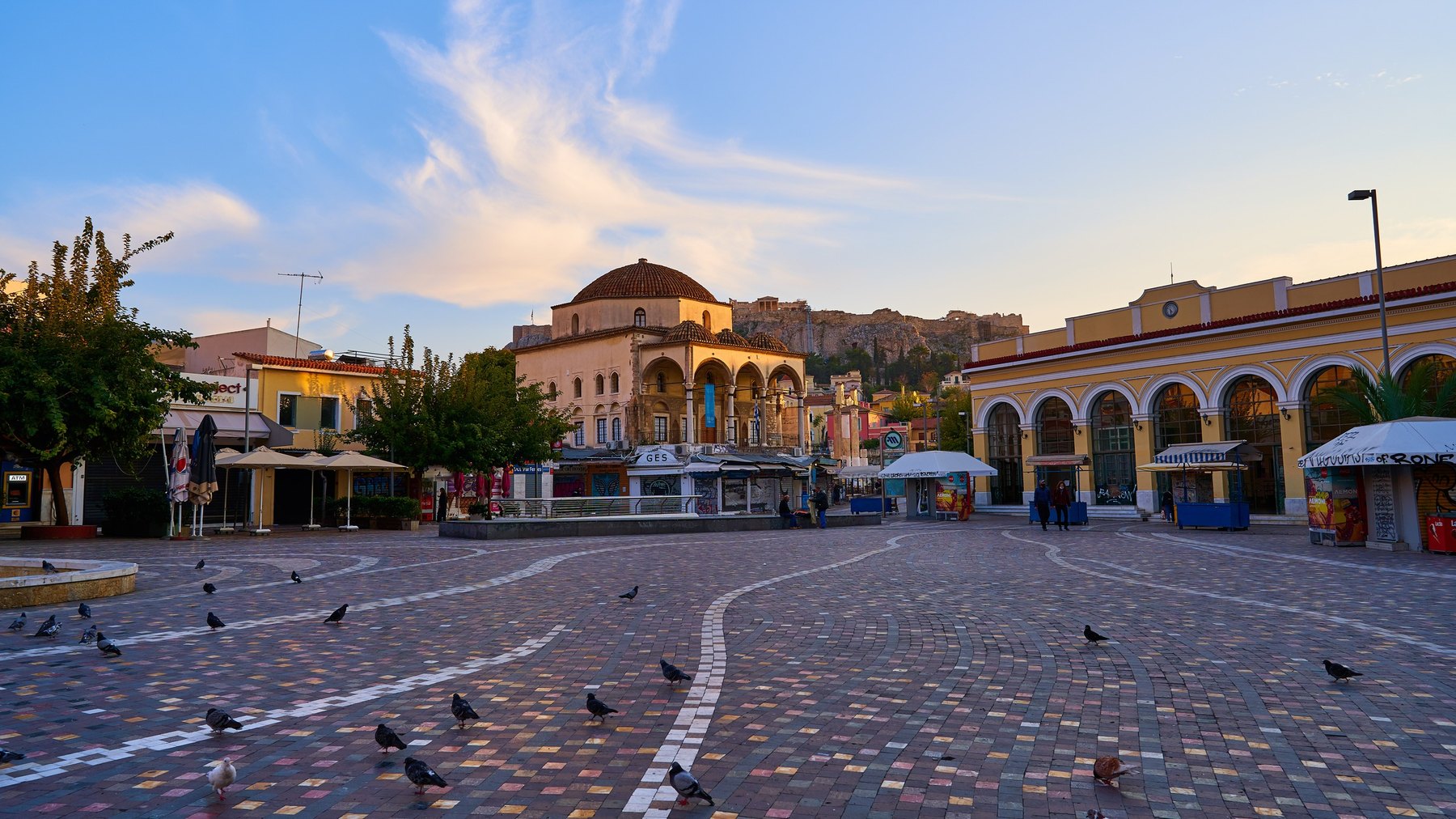 Monastiraki Square in Athens at golden hour, nearly empty, with the neoclassical ochre arcade of the 1895 metro station building, the domed Tzistarakis Mosque and the Acropolis rock behind